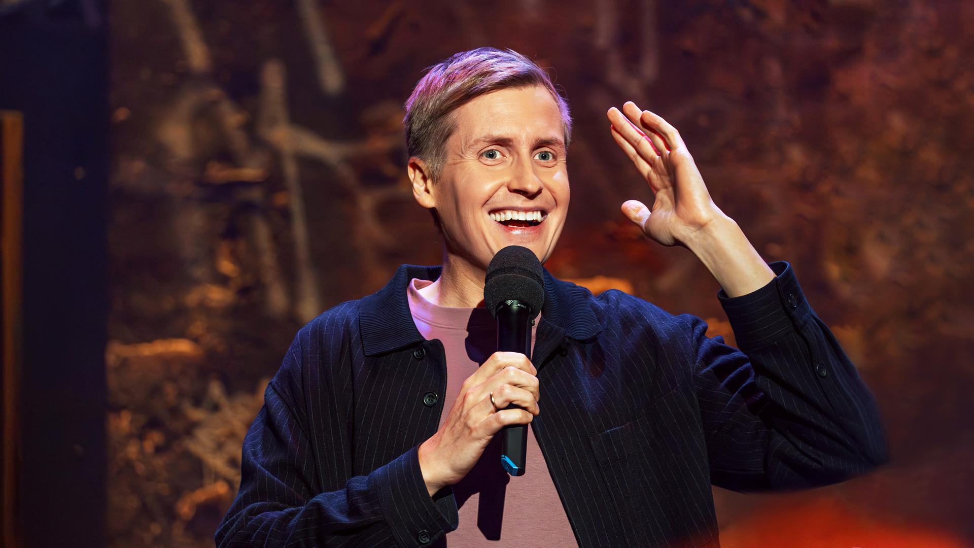 Till Reiners ist auf der rechten Bildseite zu sehen. In seiner rechten Hand hält er ein Mikrofon und hebt dazu den linken Arm auf Kopfhöhe an. Er lächelt.