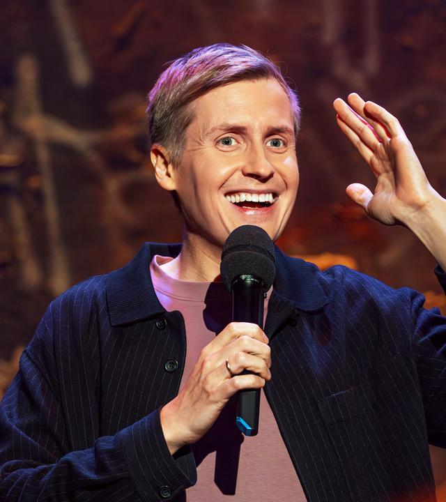 Till Reiners ist auf der rechten Bildseite zu sehen. In seiner rechten Hand hält er ein Mikrofon und hebt dazu den linken Arm auf Kopfhöhe an. Er lächelt.