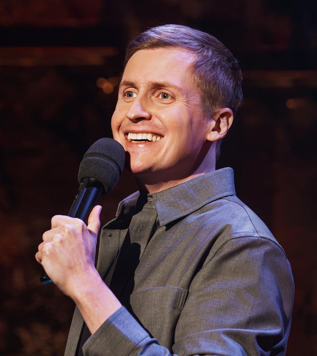 Till Reiners ist auf der rechten Bildseite zu sehen. In seiner linken Hand hält er ein Mikrofon. Der Hintergrund ist dunkel, sein Hemd ist grau.