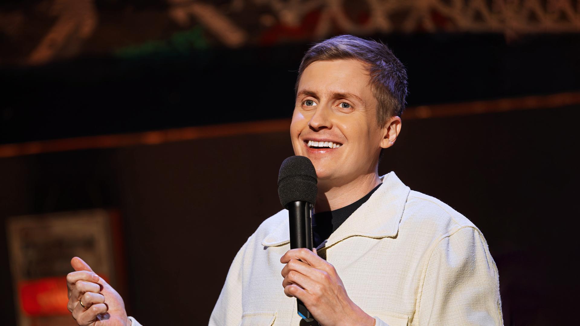 Till Reiners ist auf der rechten Bildseite zu sehen. In seiner linken Hand hält er ein Mikrofon, die rechte Hand ist nur am unteren Bildrand zu sehen. Er trägt eine offene Jacke in der Farbe beige. Er lächelt.