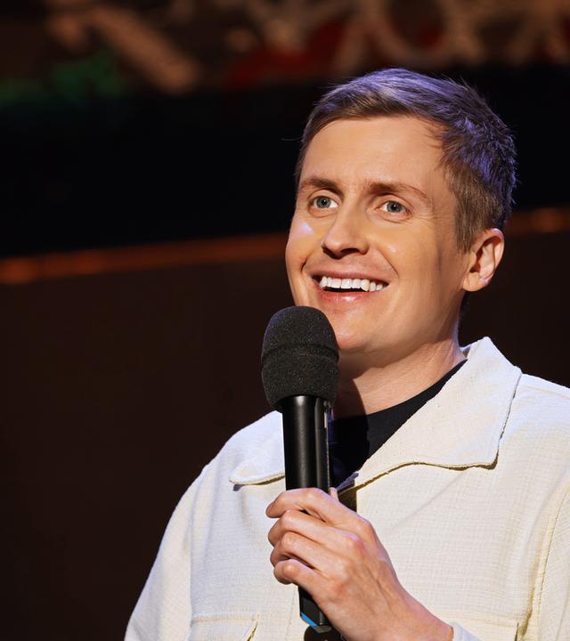 Till Reiners ist auf der rechten Bildseite zu sehen. In seiner linken Hand hält er ein Mikrofon, die rechte Hand ist nur am unteren Bildrand zu sehen. Er trägt eine offene Jacke in der Farbe beige. Er lächelt.