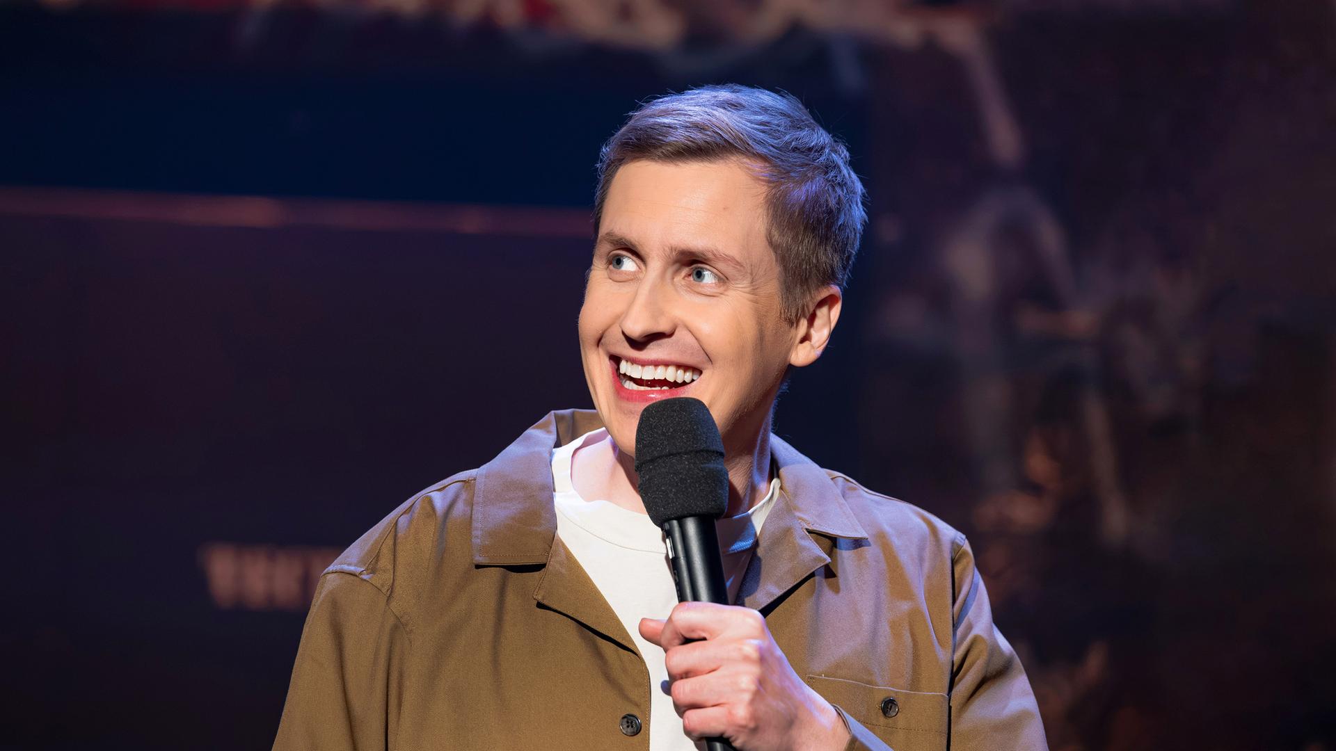 Till Reiners ist auf der rechten Bildseite zu sehen. In seiner linken Hand hält er ein Mikrofon. Er trägt eine offene Jacke in der Farbe beige. Er lächelt.