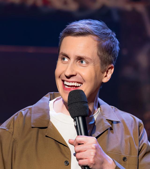 Till Reiners ist auf der rechten Bildseite zu sehen. In seiner linken Hand hält er ein Mikrofon. Er trägt eine offene Jacke in der Farbe beige. Er lächelt.