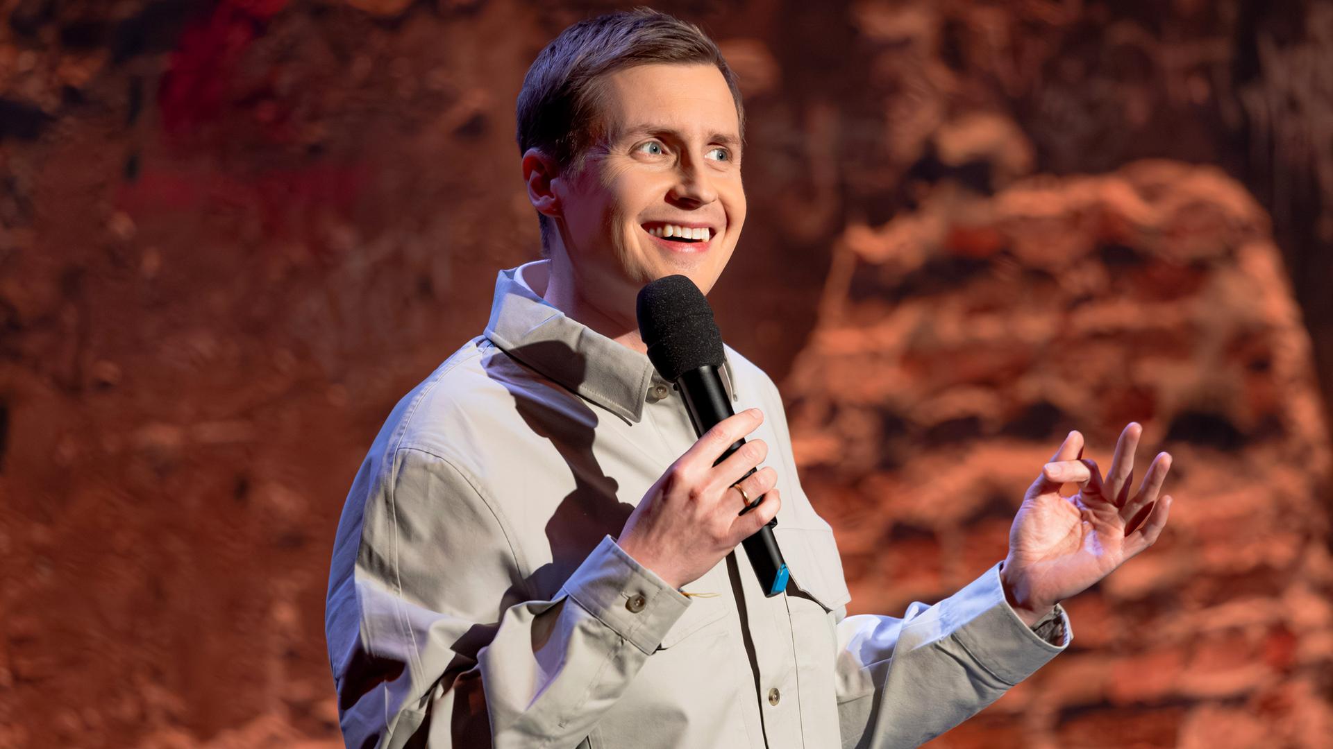 Till Reiners steht in der Mitte des Bildes. In seiner rechten Hand hält er ein Mikrofon. Er trägt ein Hemd. Till Reiners lächelt.