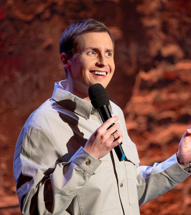 Till Reiners steht in der Mitte des Bildes. In seiner rechten Hand hält er ein Mikrofon. Er trägt ein Hemd. Till Reiners lächelt.