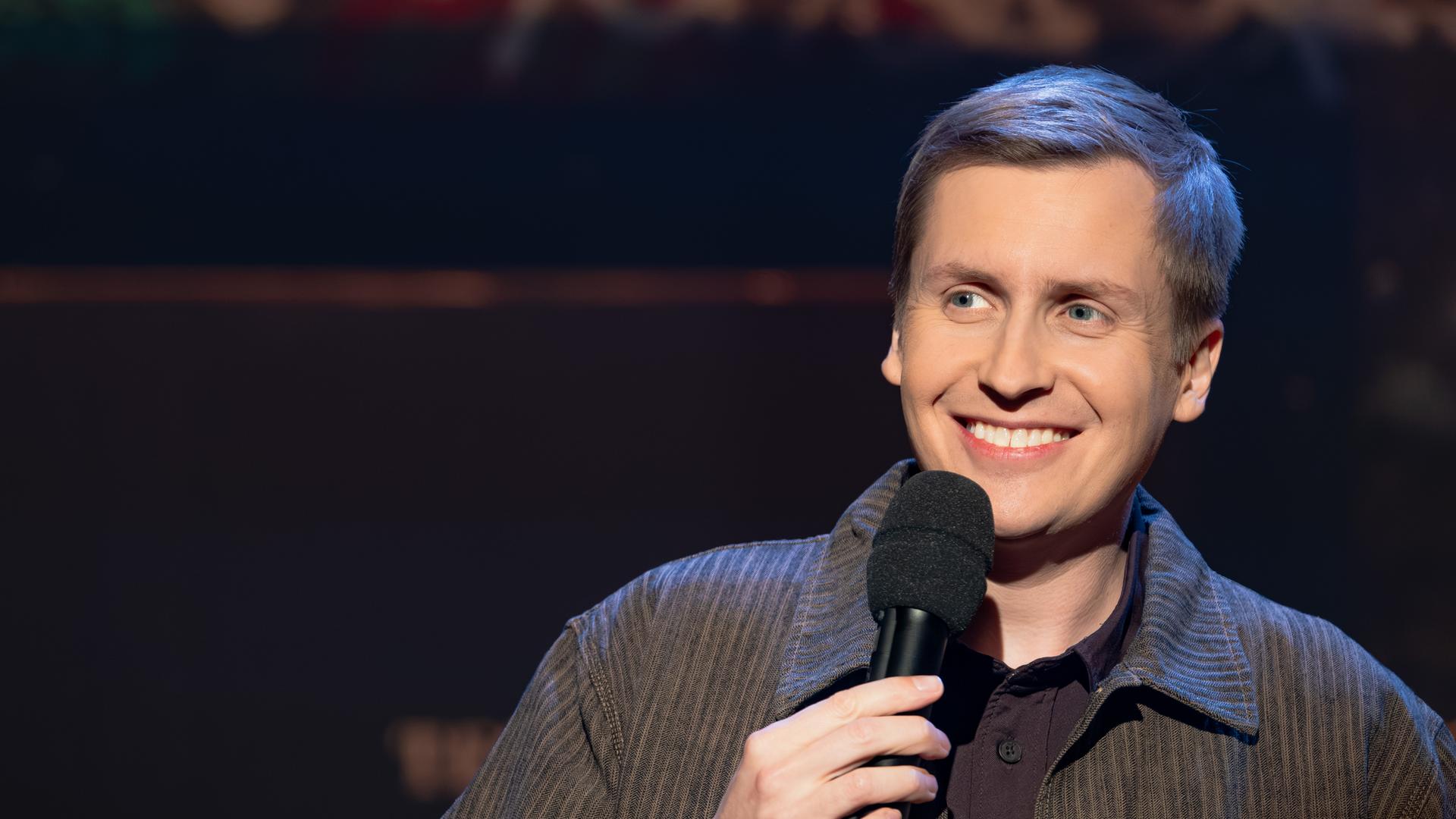 Till Reiners ist auf der rechten Seite des Bildes abgebildet. In seiner rechten Hand hält er ein Mikrofon. Reiners lächelt. Der Hintergrund ist dunkel.