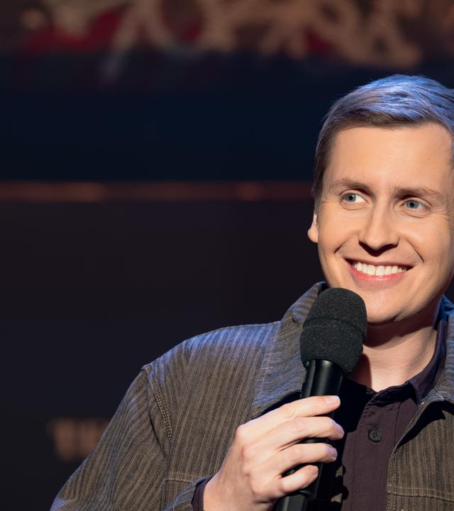 Till Reiners ist auf der rechten Seite des Bildes abgebildet. In seiner rechten Hand hält er ein Mikrofon. Reiners lächelt. Der Hintergrund ist dunkel.