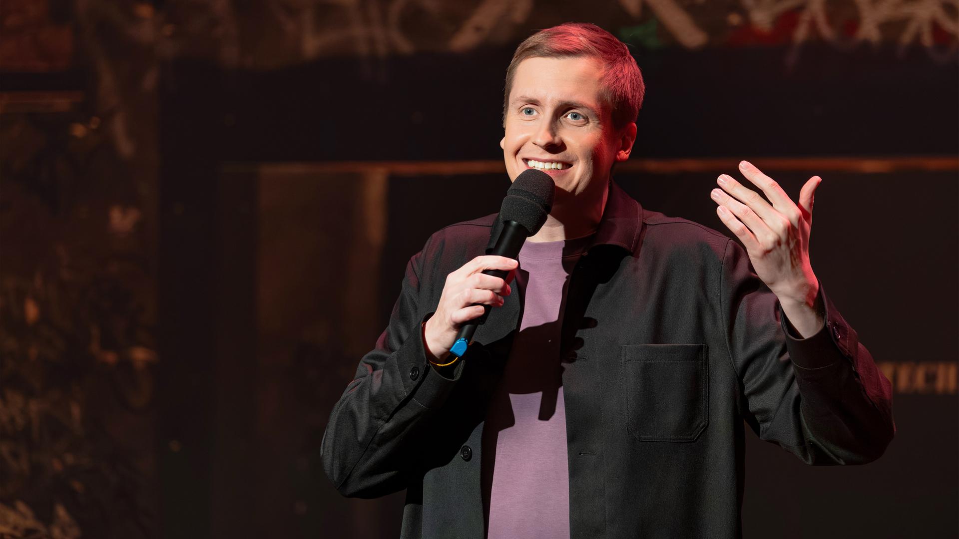 Till Reiners ist in der Mitte des Bildes abgebildet. In seiner rechten Hand hält er ein Mikrofon. Reiners lächelt. Der Hintergrund ist dunkel. Er hebt seine linke Hand. 