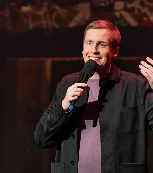 Till Reiners ist in der Mitte des Bildes abgebildet. In seiner rechten Hand hält er ein Mikrofon. Reiners lächelt. Der Hintergrund ist dunkel. Er hebt seine linke Hand. 