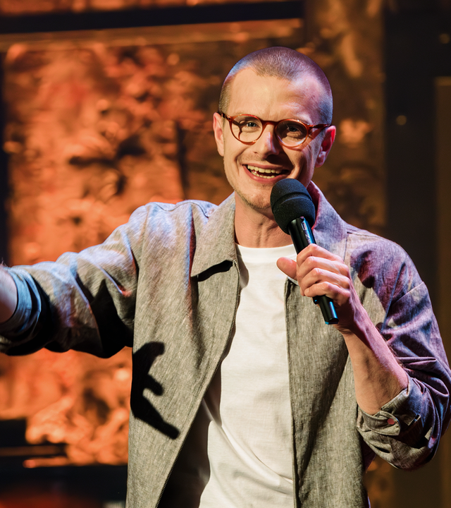 Moritz Neumeier steht in der Mitte des Bildes. In seiner linken Hand hält er ein Mikrofon. Er trägt eine Brille und ein offenes Hemd mit einem weißen T-Shirt darunter.