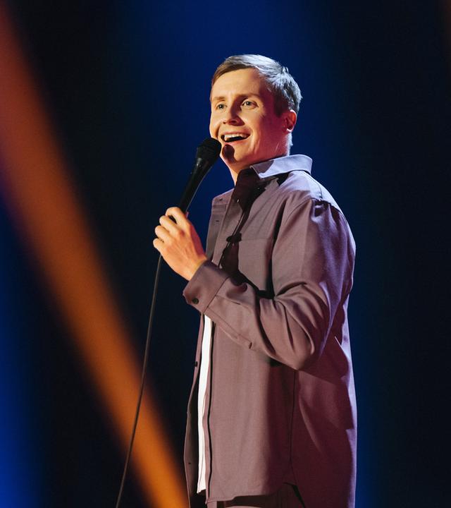 Till Reiners steht auf der rechten Bildseite. In seiner linken Hand hält er ein Mikrofon. Der Hintergrund ist blau und orange beleuchtet.