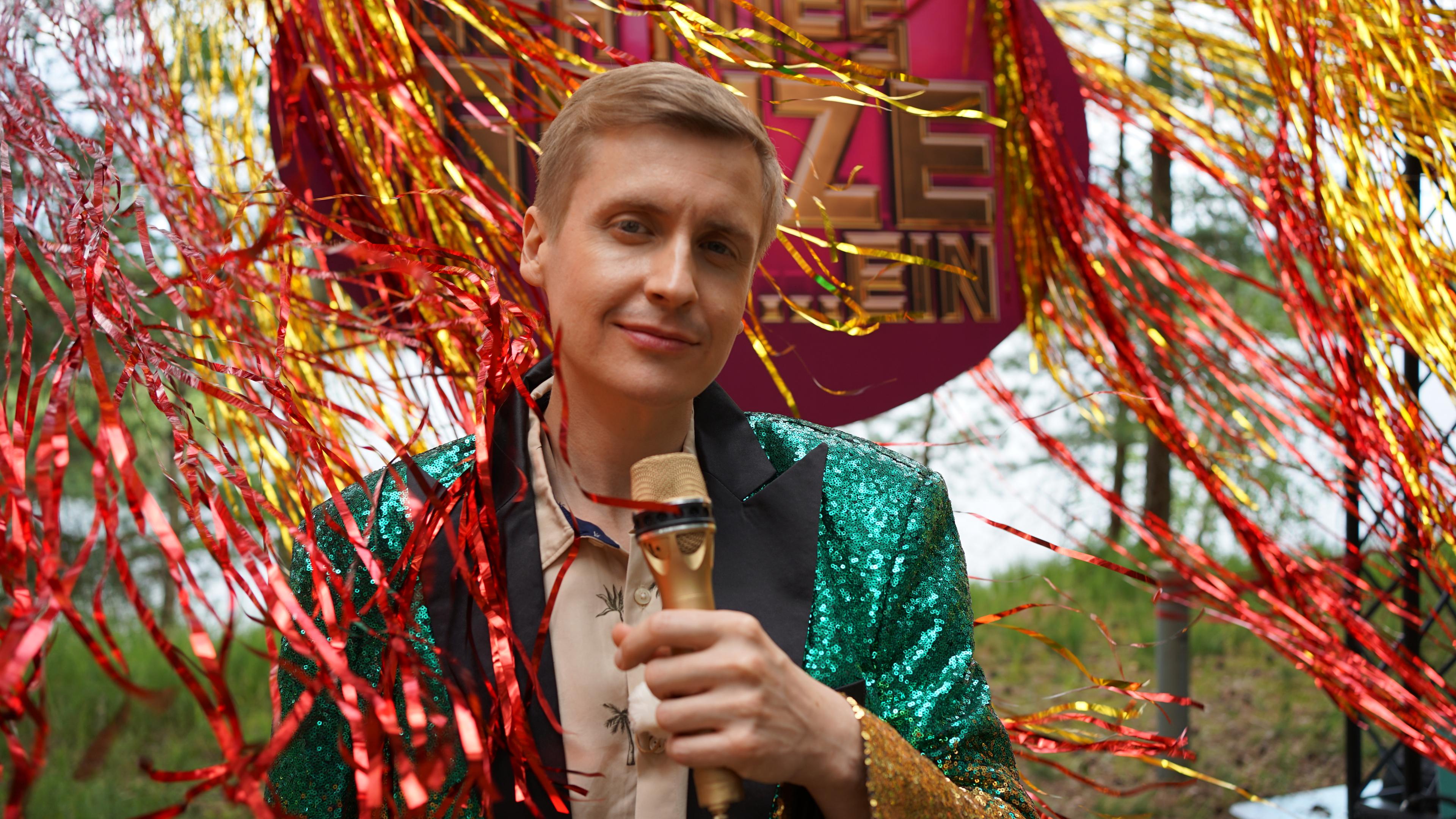 Till Reiners steht mit einem grün-schillernden Anzug und einem goldenen Mikro in der Hand draußen inmitten von Lametta, das rot und gold glänzt 
