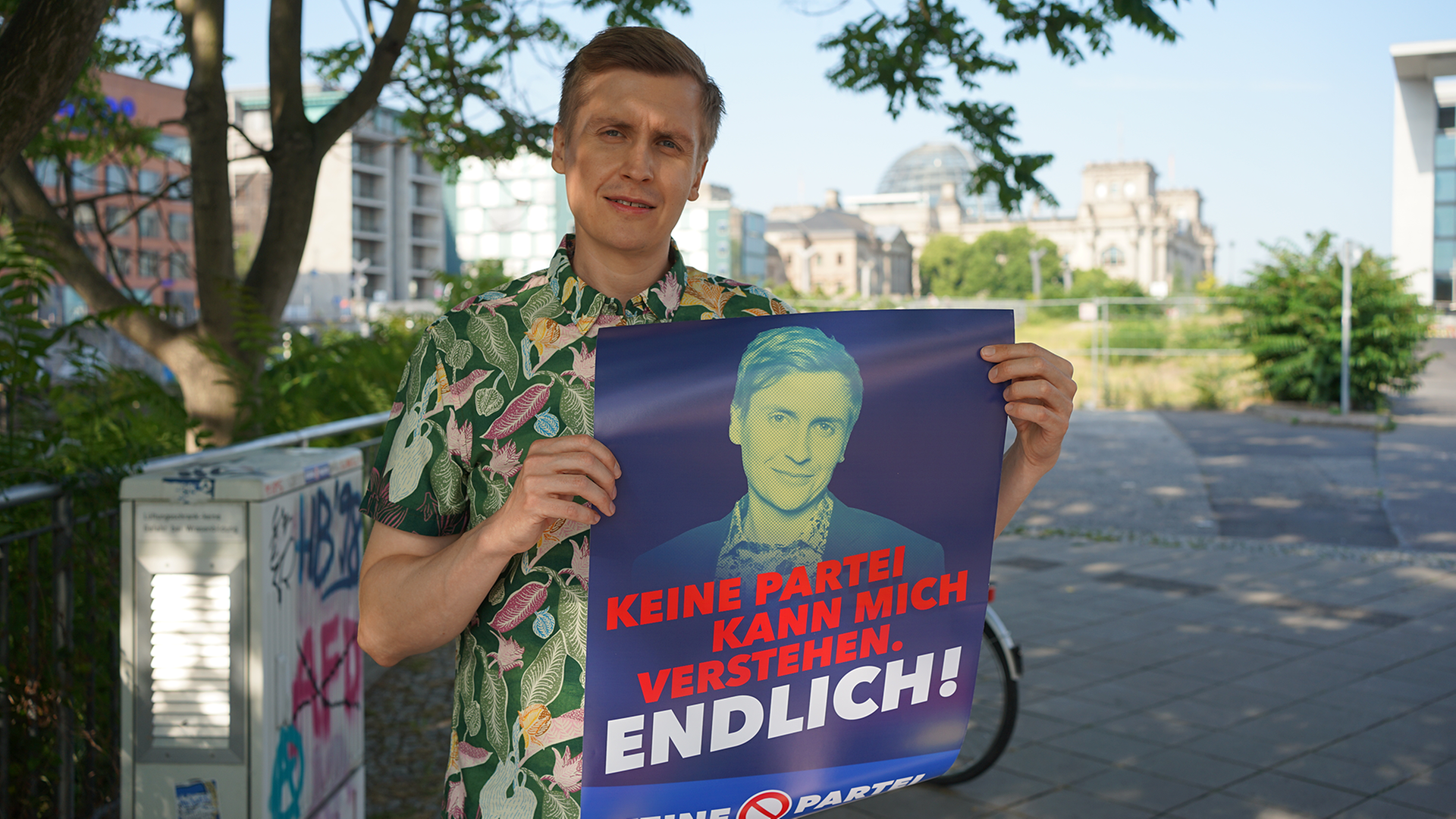 Till Reiners steht in einem Hawaihemd draußen und hält ein Wahlplakat in der Hand, auf dem zu lesen ist: Keine Partei kann mich verstehen. Endlich