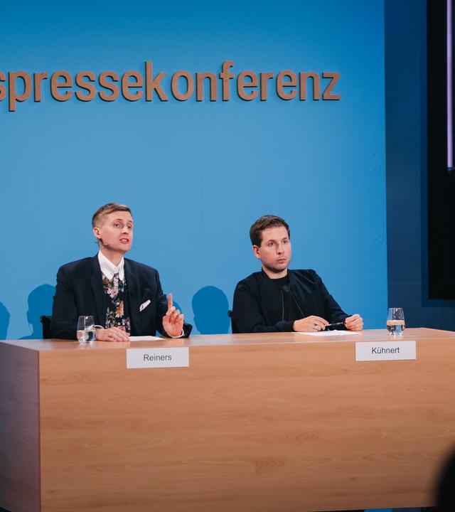 Till Reiners und Kevin Kühnert vor einer nachgebauten Wand der Bundespressekonferenz.