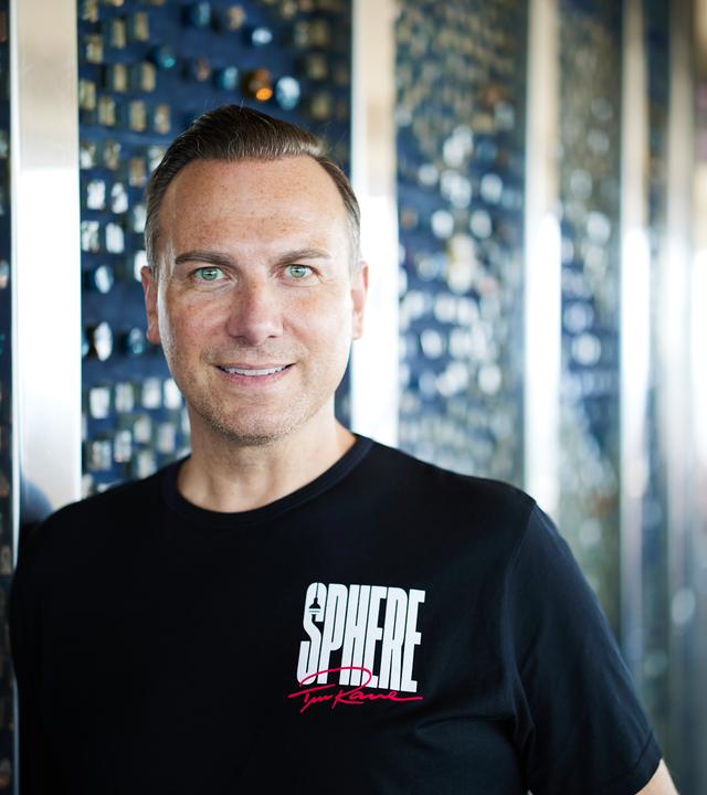 Porträtfoto eines Mannes mit ganz kurzen mittelblonden Haaren und blauen Augen, der ein schwarzes T-Shirt mit der weißen Aufschrift Sphere trägt.