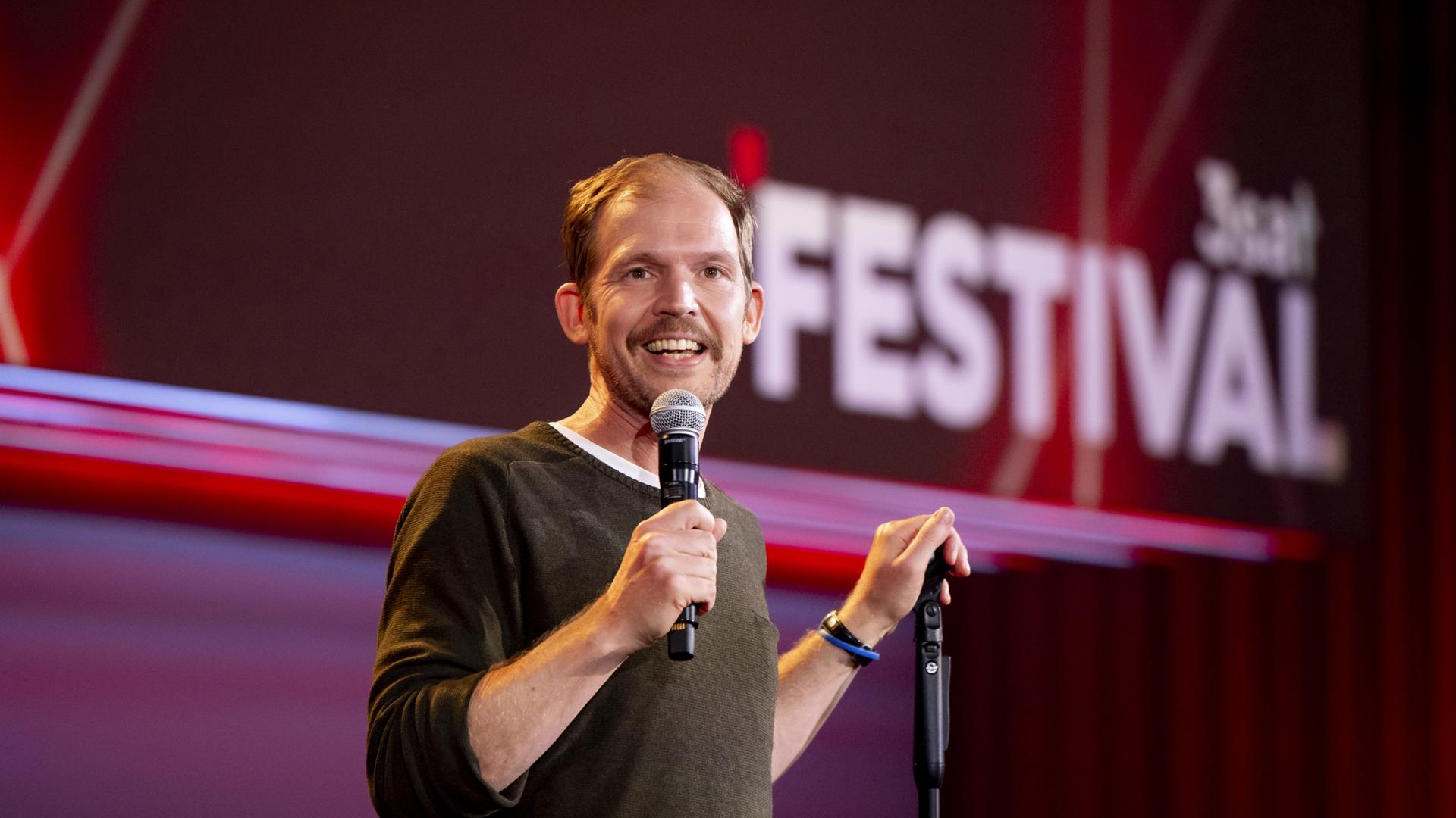 Timo Wopp steht in der Mitte des Bildes. Mit seiner linken Hand hält er einen Mikrofonständer. In seiner rechten Hand hält er ein Mikrofon. Der Hintergrund ist rot beleuchtet.