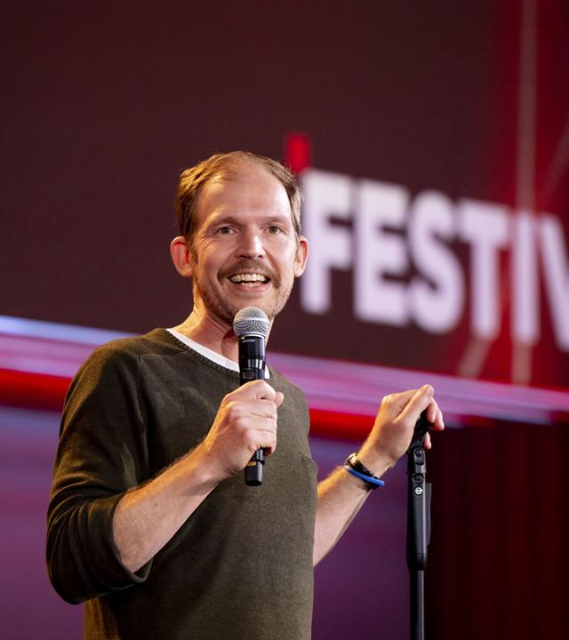 Timo Wopp steht in der Mitte des Bildes. Mit seiner linken Hand hält er einen Mikrofonständer. In seiner rechten Hand hält er ein Mikrofon. Der Hintergrund ist rot beleuchtet.