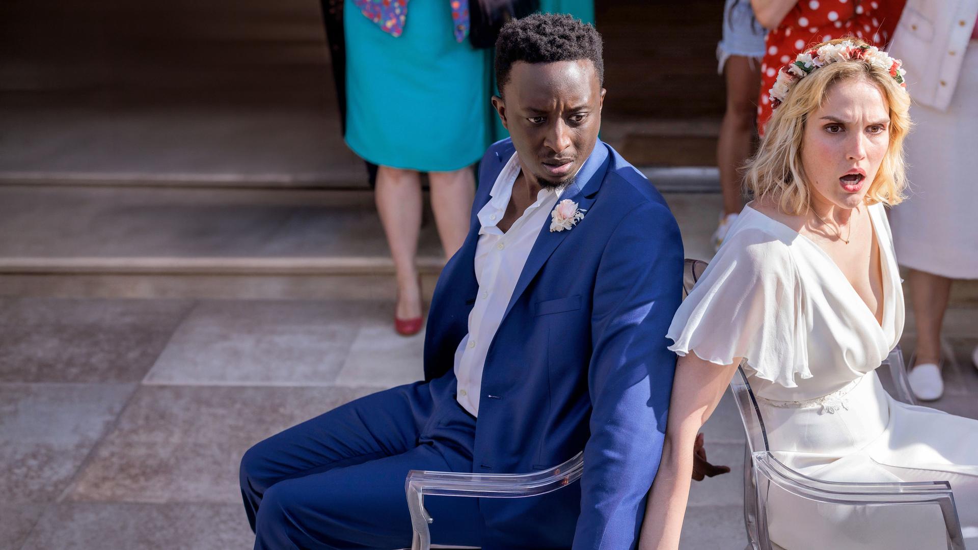 "Tiny Wedding - Unsere mega kleine Hochzeit": Halbnahe, leichte Aufsicht: Max (Ahmed Sylla) und Lou (Camille Lou) sitzen in ihren Hochzeitsoutfits mit schockierten Gesichtern Rücken an Rücken an Stühle gefesselt vor einer Feiergesellschaft. Diese ist nur hüftabwärts im Bild zu sehen.