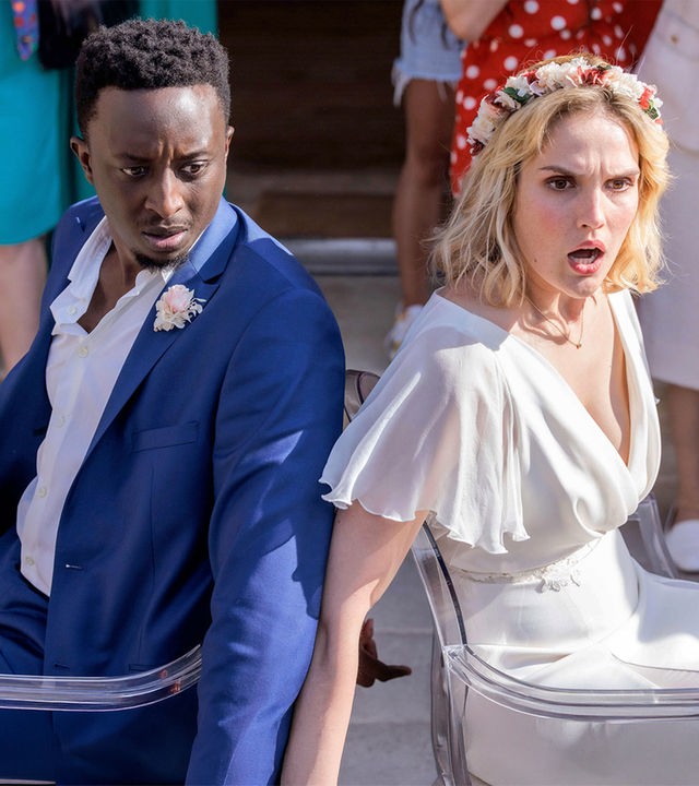 "Tiny Wedding - Unsere mega kleine Hochzeit": Halbnahe, leichte Aufsicht: Max (Ahmed Sylla) und Lou (Camille Lou) sitzen in ihren Hochzeitsoutfits mit schockierten Gesichtern Rücken an Rücken an Stühle gefesselt vor einer Feiergesellschaft. Diese ist nur hüftabwärts im Bild zu sehen.