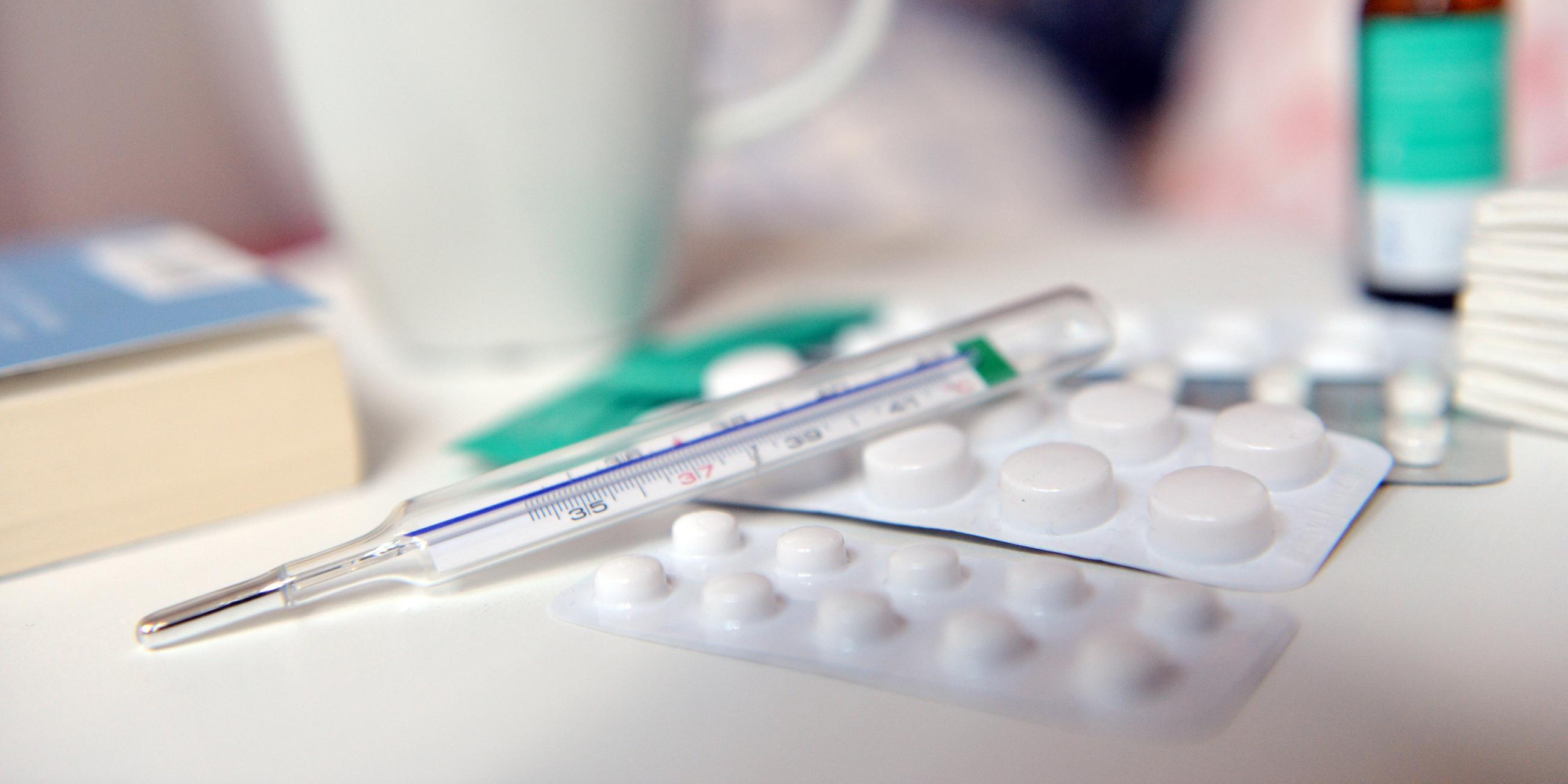 Auf dem Foto sieht man im Vordergrund zwei weiße Tablettenpackungen mit einem Fieberthermometer Glas. Daneben liegt ein blaues Buch und im Hintergrund eine weiße Tasse. 