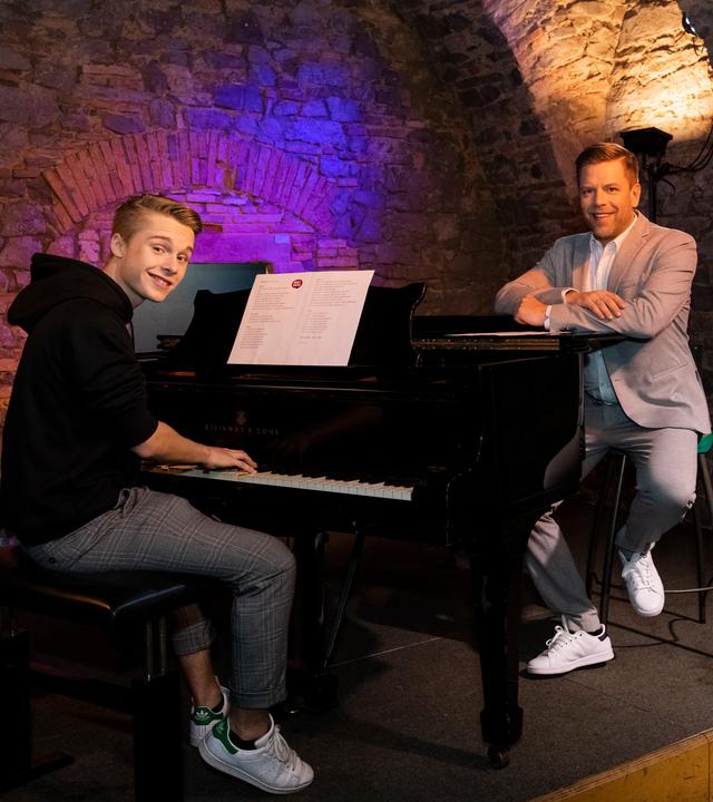 "Dein Song - Tipps von den Paten - Folge 13":  Lion sitzt am Flügel im Jazzkeller. Ihm gegenüber sitzt sein Pate Tom Gaebel mit den Armen auf dem Flügel abgelegt und lächelt in die Kamera.