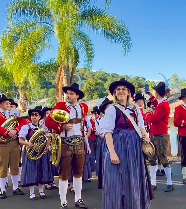 Das Bild zeigt eine Blasmusik-Kapelle in einer festlichen Umgebung. Im Vordergrund stehen mehrere Musiker in traditionellen Trachten mit roten Jacken und breiten Hüten, die mit Federn geschmückt sind. Einige von ihnen halten Instrumente wie Tuba und Trompete. Die meisten tragen Hosen in Erdtönen, kombiniert mit weißen Socken.   Eine auffällig gekleidete Person mit Brille steht in der Mitte des Bildes und hat eine auffallende Schärpe umgelegt. Hinter den Musikern sieht man eine Reihe von Palmen, die auf das warme Klima hindeuten. Der Himmel ist strahlend blau, und im Hintergrund sind einige Gebäude, die möglicherweise zur festlichen Veranstaltung oder zur Gemeinde gehören. Die gesamte Szene vermittelt ein Gefühl von Gemeinschaft und kulturellem Erbe.