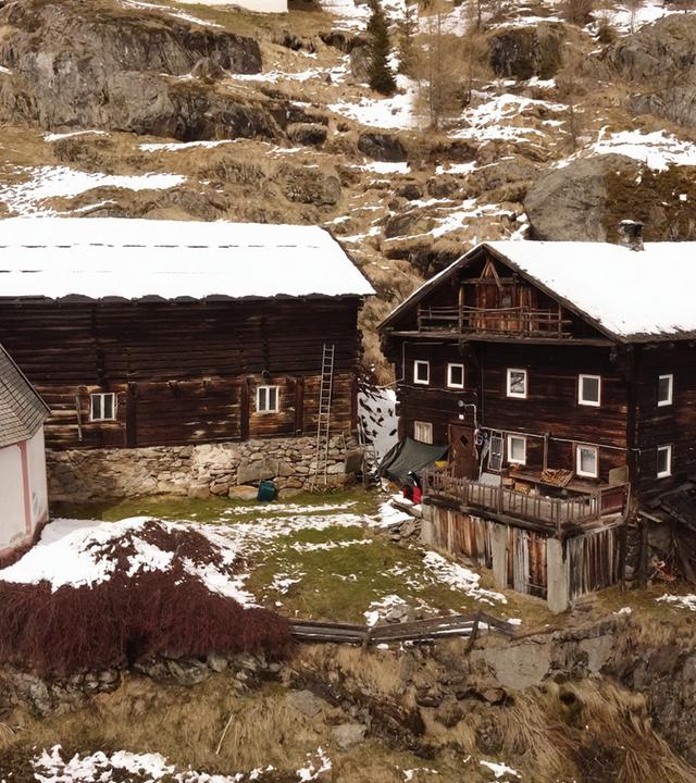 Das Bild zeigt eine ländliche Szenerie in Tirol. Im Vordergrund sind zwei traditionelle Bauernhäuser zu sehen. Das linke Gebäude hat eine helle Fassade mit einem runden, spitzen Dach und einer kleinen, ornamente Stube. An der Wand befindet sich eine dekorative Malerei. Das rechte Gebäude ist aus dunklem Holz konstruiert, hat mehrere Fenster und einen Balkon. Die Dächer beider Häuser sind teilweise mit Schnee bedeckt.  Im Hintergrund ist eine hügelige, felsige Landschaft sichtbar, die ebenfalls mit Schnee bedeckt ist. Der Boden ist größtenteils braun und trocken, und einige Grasflächen sind sichtbar. Eine Leiter lehnt an dem linken Haus, und bis auf einige grasbewachsenen Flächen sind keine weiteren Pflanzen vorhanden. Es ist ein klarer, kalter Tag, was durch die Schneebedeckung und die winterliche Umgebung suggeriert wird.