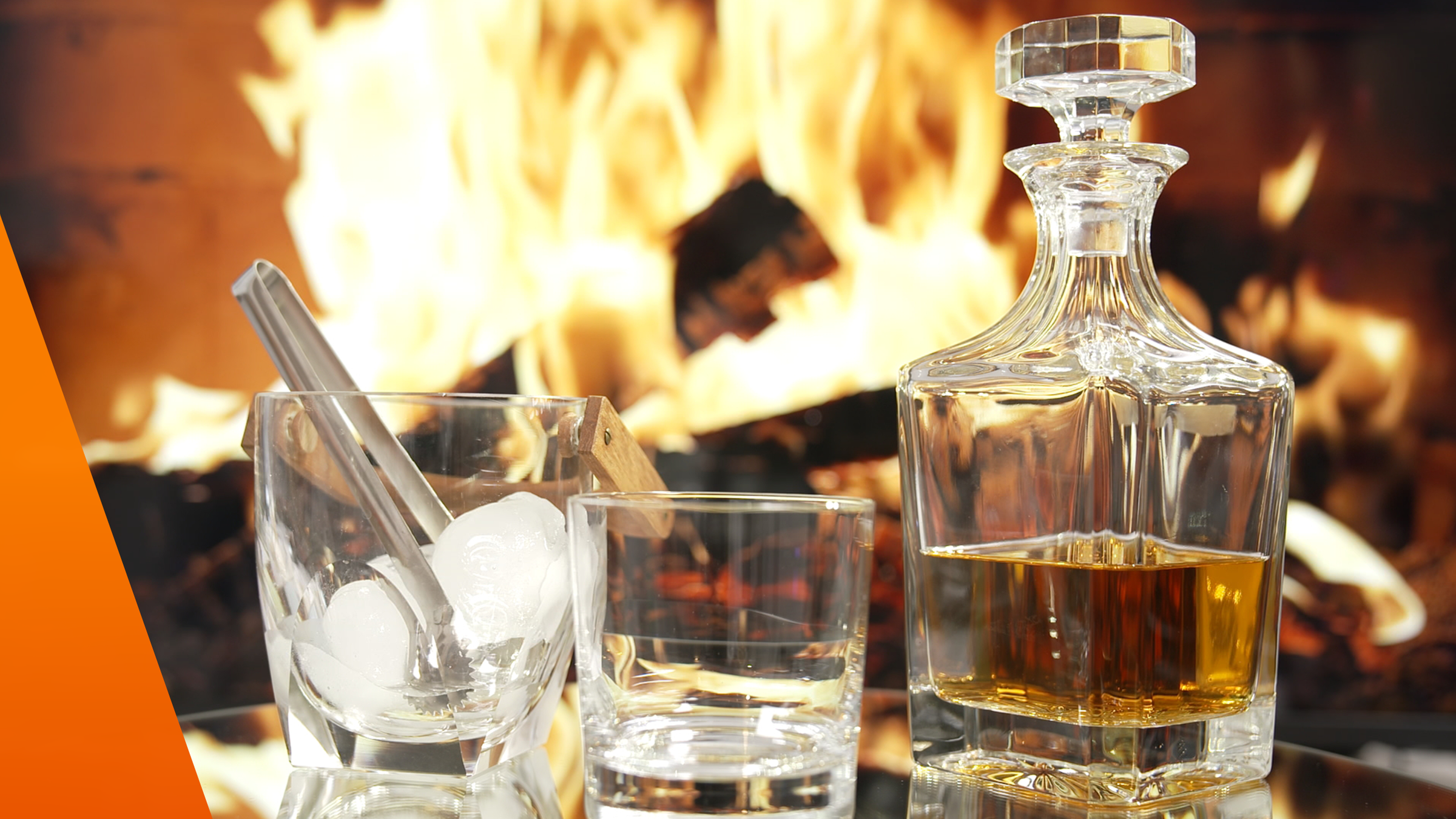 Whiskey in Karaffe mit zwei Gläsern. In einem Glas sind Eiswürfel. Im Hintergrund lodert ein Kaminfeuer.