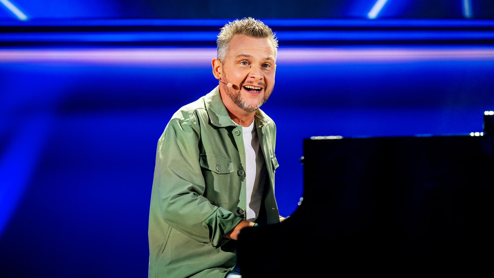 Kabarettist sitzt am Flügel in der Mitte des Bildes. Er trägt ein offenes grünes Hemd. Darunter trägt er ein weißes Shirt. Der Hintergrund ist blau beleuchtet.