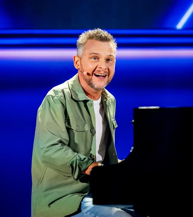 Kabarettist sitzt am Flügel in der Mitte des Bildes. Er trägt ein offenes grünes Hemd. Darunter trägt er ein weißes Shirt. Der Hintergrund ist blau beleuchtet.