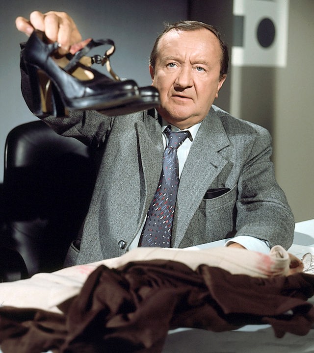 "Der Alte - Toccata und Fuge": Hauptkommissar Köster (Siegried Lowitz) sitzt an einem mit Kleider belegten Schreibtisch und einem Telefon, ganz rechts im Bild. Eine Lesebrille liegt auch noch auf dem Tisch. Er hält ein paar schwarze Damenschuhe in die Kamera.