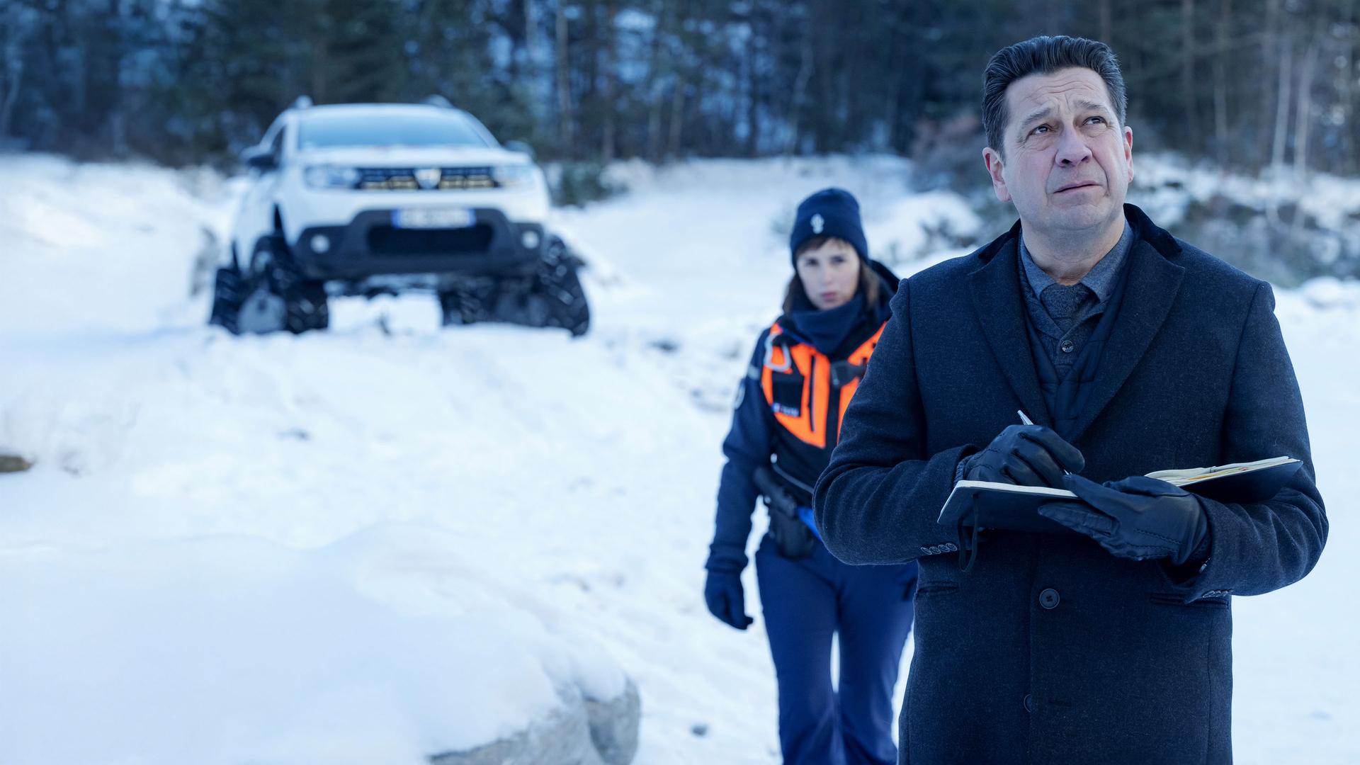 "Tod auf dem Gipfel": Halbnahe: Andréas (Laurent Gerra) steht mit Stift und aufgeschlagenem Notizbuch in einer verschneiten Landschaft und blickt konzentriert nach oben. Hinter Andréas läuft Constance (Clémentine Poidatz) in einer Warnweste auf ihn zu. Im Hintergrund steht ein weißer Jeep am Waldrand geparkt.
