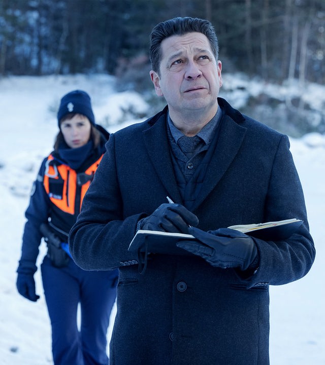 "Tod auf dem Gipfel": Halbnahe: Andréas (Laurent Gerra) steht mit Stift und aufgeschlagenem Notizbuch in einer verschneiten Landschaft und blickt konzentriert nach oben. Hinter Andréas läuft Constance (Clémentine Poidatz) in einer Warnweste auf ihn zu. Im Hintergrund steht ein weißer Jeep am Waldrand geparkt.