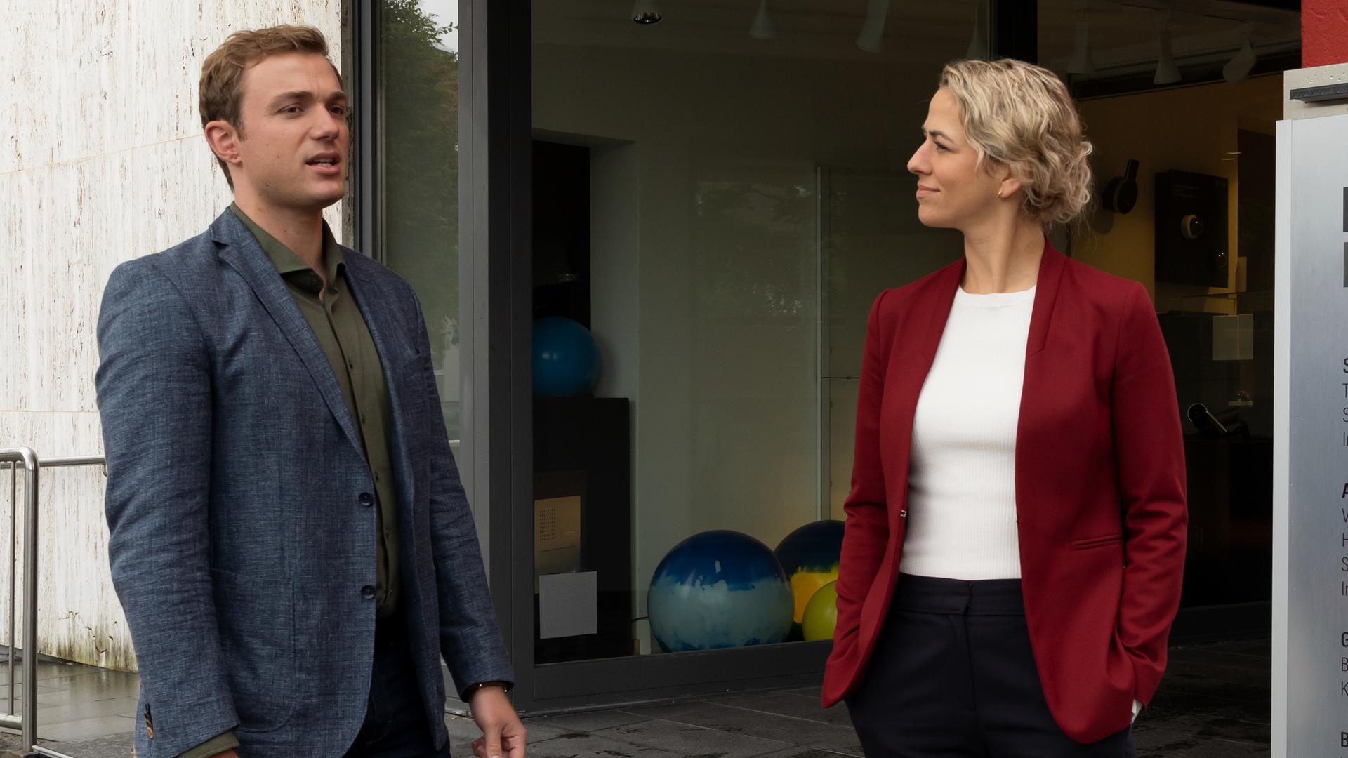 Die Kommissare Thomas (Moritz von Zeddelmann) und Laura Schmidt (Marija Kovčo) stehen am Treppenaufgang des Geschäfts "Reinsbach Elektrohandel". Auf dem Schild rechts von Laura Schmidt sind neben dem Geschäftsnamen auch die Dienstleistungen aufgeführt.