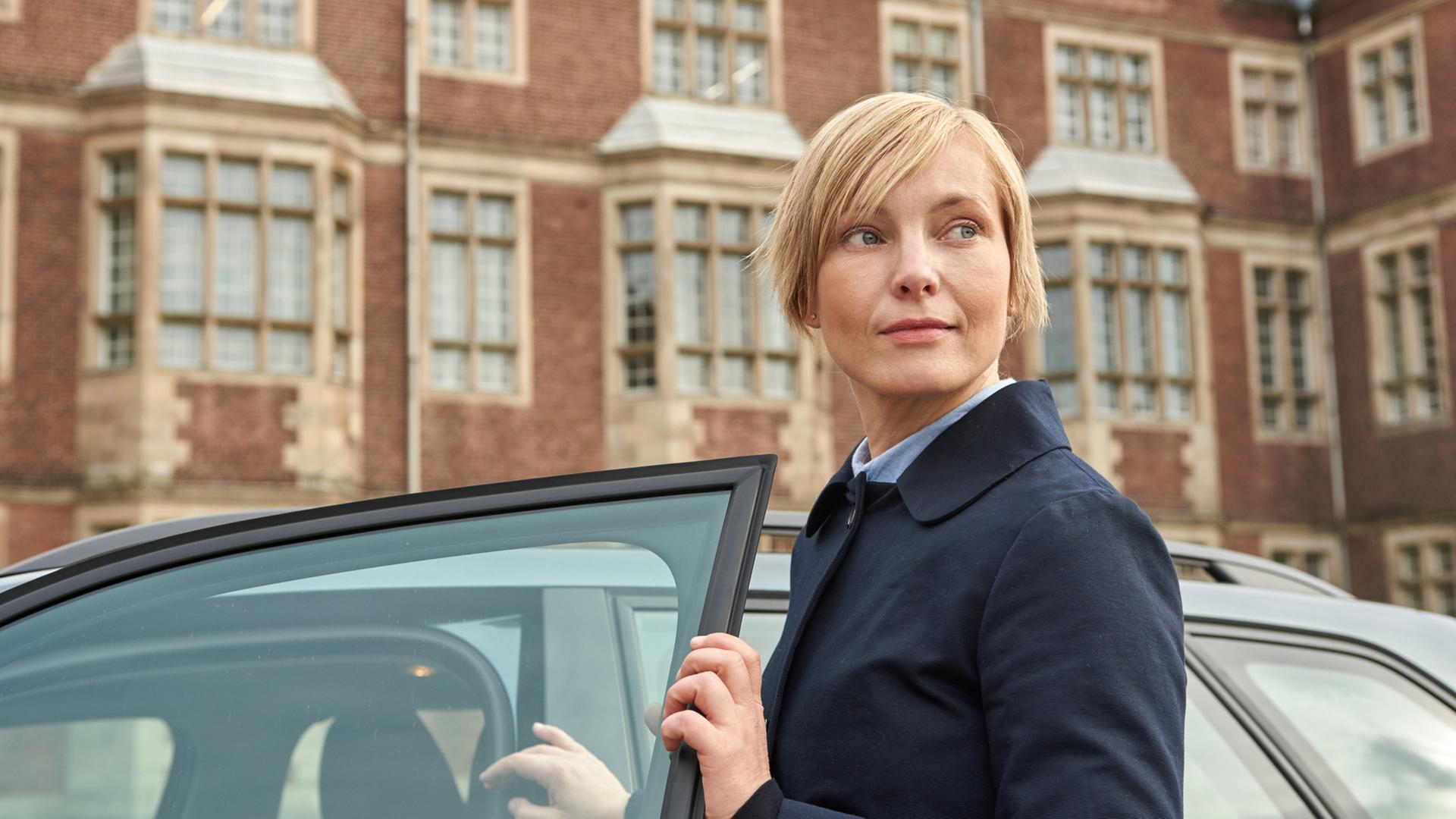 "Tod im Internat": Lehrerin Karla Parker (Mitte rechts) ist gerade aus einem Auto ausgestiegen und schaut nun, mit der Autotür in der Hand, in die Ferne. Im Hintergrund ist die Hausfassade des Elite-Internats Erlengrund zu sehen. Tatsächlich ist sie die verdeckte Ermittlerin Isabell Moosbach (Nadja Uhl).