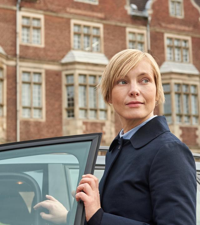 "Tod im Internat": Lehrerin Karla Parker (Mitte rechts) ist gerade aus einem Auto ausgestiegen und schaut nun, mit der Autotür in der Hand, in die Ferne. Im Hintergrund ist die Hausfassade des Elite-Internats Erlengrund zu sehen. Tatsächlich ist sie die verdeckte Ermittlerin Isabell Moosbach (Nadja Uhl).