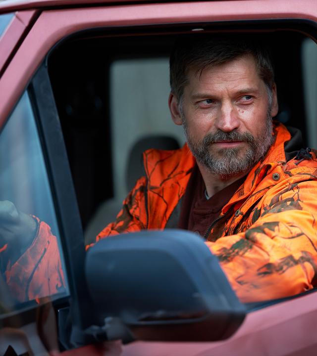 Ein bärtiger Mann mit kurzer Frisur sitzt am Steuer eines Autos und trägt eine orange Tarnjacke. Er blickt ernst aus dem Fenster.