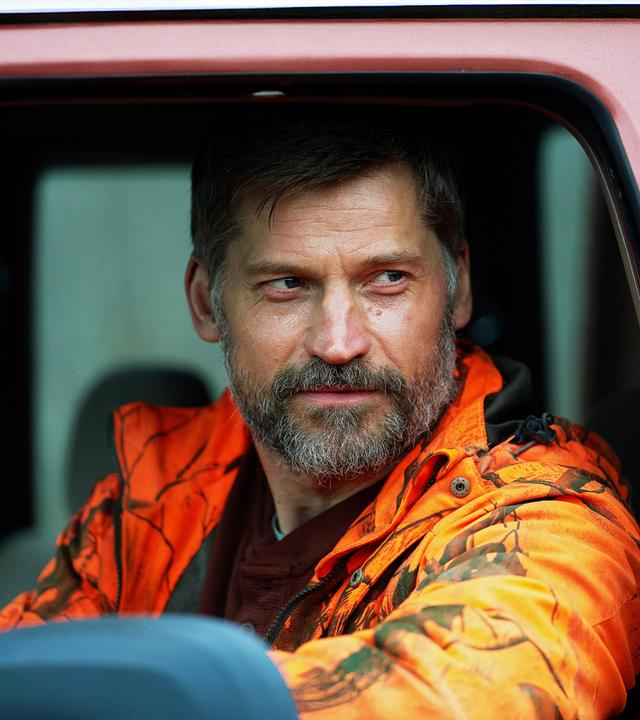 Ein bärtiger Mann mit kurzer Frisur sitzt am Steuer eines Autos und trägt eine orange Tarnjacke. Er blickt ernst aus dem Fenster.