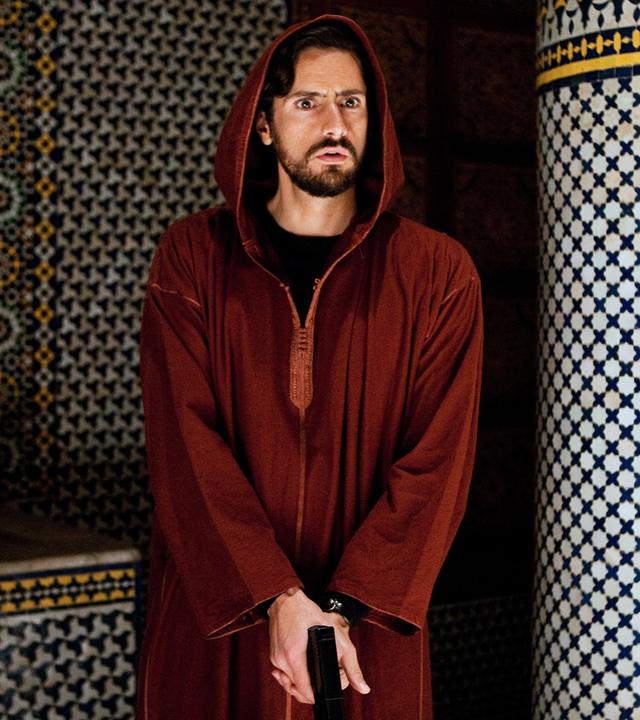 Kommissar Falcón (Juan Diego Botto) steht mit der Pistole in der Hand neben einer Säule und beobachtet entgeistert etwas.