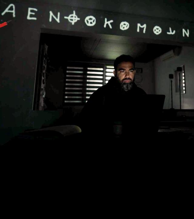 Rechts ein Mann vor dem Bildschirm seines Computers, über ihm auf die Wand projeziert Teile des Codes, den der Zodiackiller benutzt hat. Links das Phantombild des Zodiackillers auf einem Stapel alter Bilder, dahinter in rot sein Zeichen