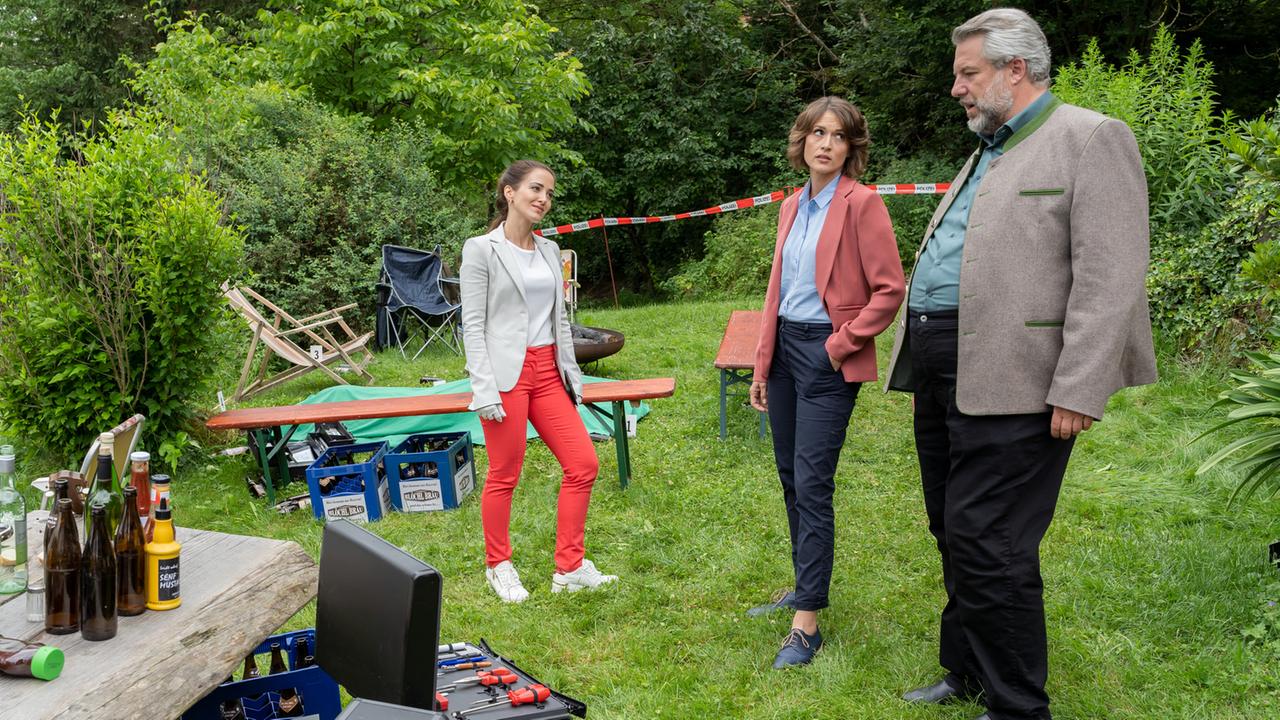 "Die Rosenheim-Cops - Tödliches Wiedersehen": Rechtsmedizinerin Atay (Sevda Polat), Kommissarin Andresen (Sophie Melbinger) und Kommissar Stadler (Dieter Fischer) stehen auf einer Wiese, die von Büschen und Bäumen umsäumt ist. Im Hintergrund sind Sitzgelegenheiten und Bierkästen zu sehen, im Vordergrund Bierflaschen auf einem Holztisch sowie die Arbeitskoffer der KTU.