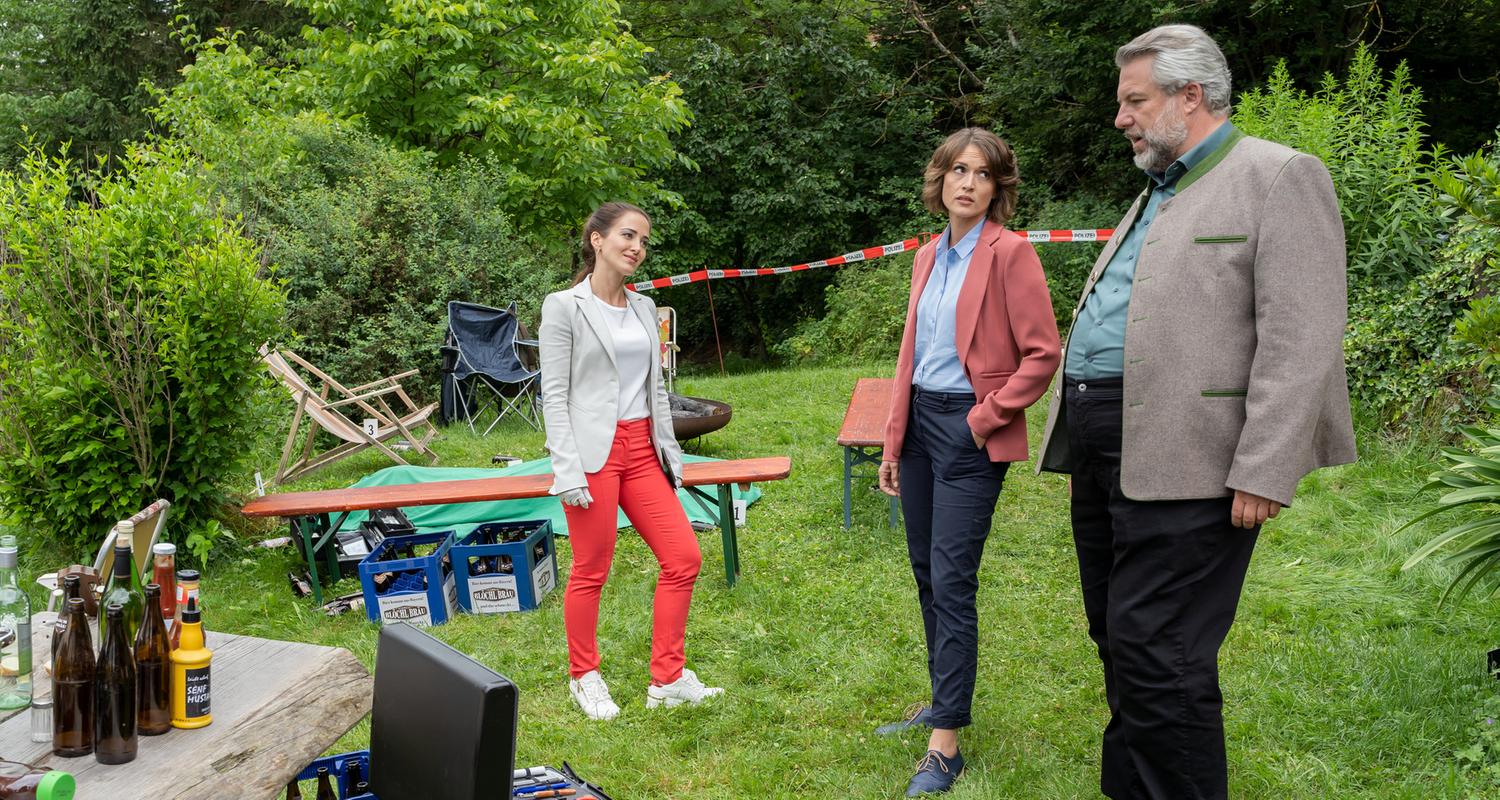 "Die Rosenheim-Cops - Tödliches Wiedersehen": Rechtsmedizinerin Atay (Sevda Polat), Kommissarin Andresen (Sophie Melbinger) und Kommissar Stadler (Dieter Fischer) stehen auf einer Wiese, die von Büschen und Bäumen umsäumt ist. Im Hintergrund sind Sitzgelegenheiten und Bierkästen zu sehen, im Vordergrund Bierflaschen auf einem Holztisch sowie die Arbeitskoffer der KTU.