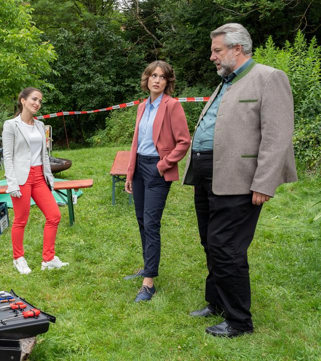 "Die Rosenheim-Cops - Tödliches Wiedersehen": Rechtsmedizinerin Atay (Sevda Polat), Kommissarin Andresen (Sophie Melbinger) und Kommissar Stadler (Dieter Fischer) stehen auf einer Wiese, die von Büschen und Bäumen umsäumt ist. Im Hintergrund sind Sitzgelegenheiten und Bierkästen zu sehen, im Vordergrund Bierflaschen auf einem Holztisch sowie die Arbeitskoffer der KTU.