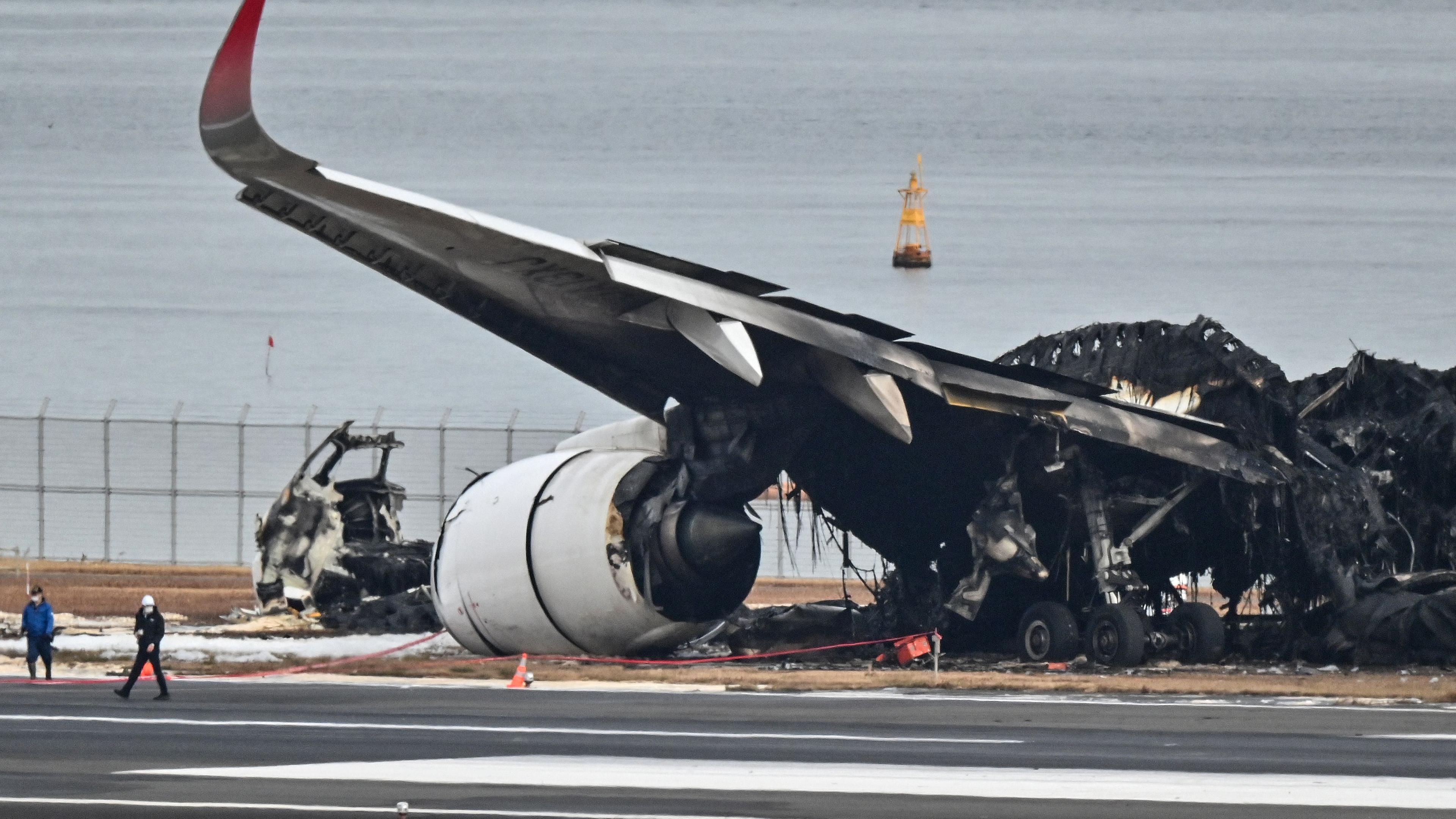 Beamte betrachten das verbrannte Wrack eines Passagierflugzeugs der Japan Airlines  auf dem Rollfeld des Tokyo International Airport in Haneda in Tokio