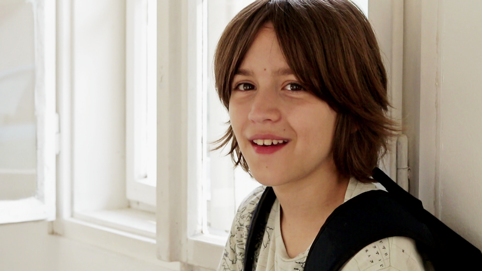 Toma (rechts im Bild) steht an einem geöffneten Fenster. Die Sonne scheint herein. Er trägt einen schwarzen Rucksack und schaut lächelnd in die Kamera.