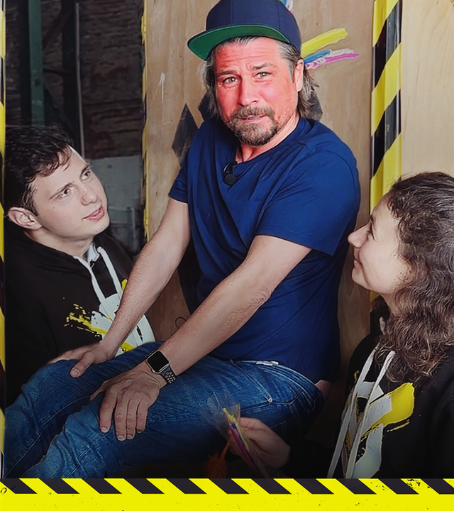 Tommy klemmt zwischen zwei Wänden. Cem und Maja stehen jeweils an seiner Seite.