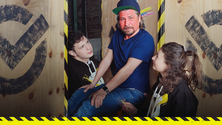 Tommy klemmt zwischen zwei Wänden. Cem und Maja stehen jeweils an seiner Seite.