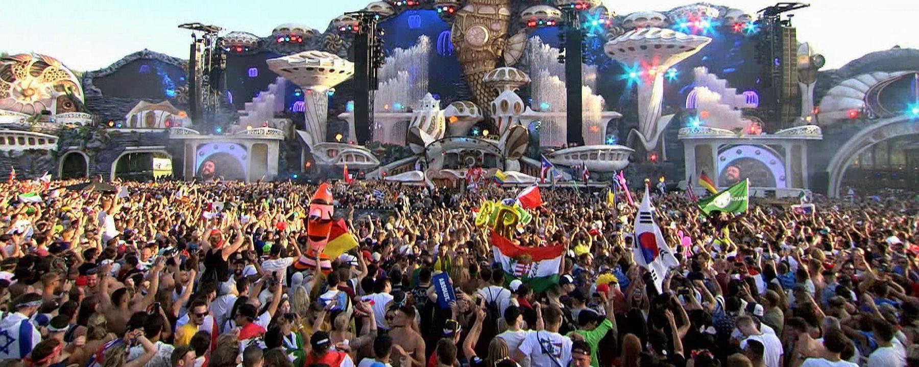 "Tomorrowland Festival - Techno, Kult und Kommerz": Hunderte Festivalbesucher feiern vor der Hauptbühne des Festivals, auf der ein DJ auftritt, und schwenken Flaggen.