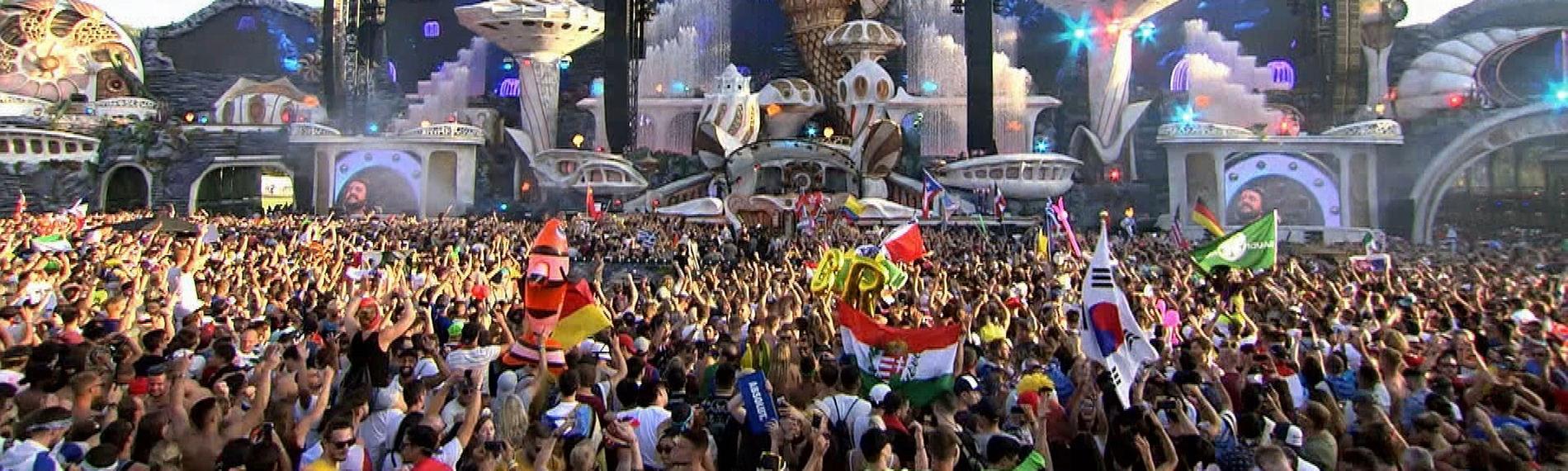 "Tomorrowland Festival - Techno, Kult und Kommerz": Hunderte Festivalbesucher feiern vor der Hauptbühne des Festivals, auf der ein DJ auftritt, und schwenken Flaggen.