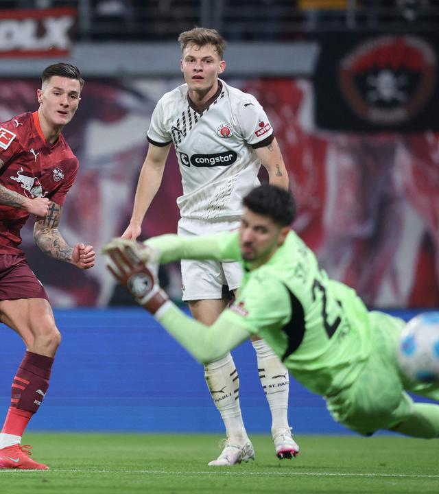 Leipzigs Stürmer (30) Benjamin Sesko (L) erzielt den Führungstreffer gegen St. Paulis Torhüter (22) Nikola Vasilj und St. Paulis Verteidiger (04) David Nemeth in Aktion.